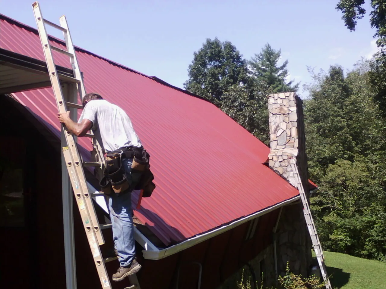 Roofer on a ladder beside a red metal roof and stone chimney — Soffit & Fascia Repair by Roofing Brothers in Hesston